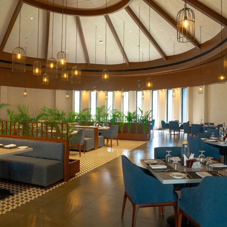 A spacious, elegant dining area with teal chairs, round tables, and a high domed ceiling with natural light at Ananta Spa and Resort, Ajabgarh.