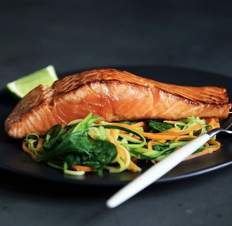 A plated dish of grilled salmon served on a bed of sautéed greens and noodles, with a lime wedge on the side.