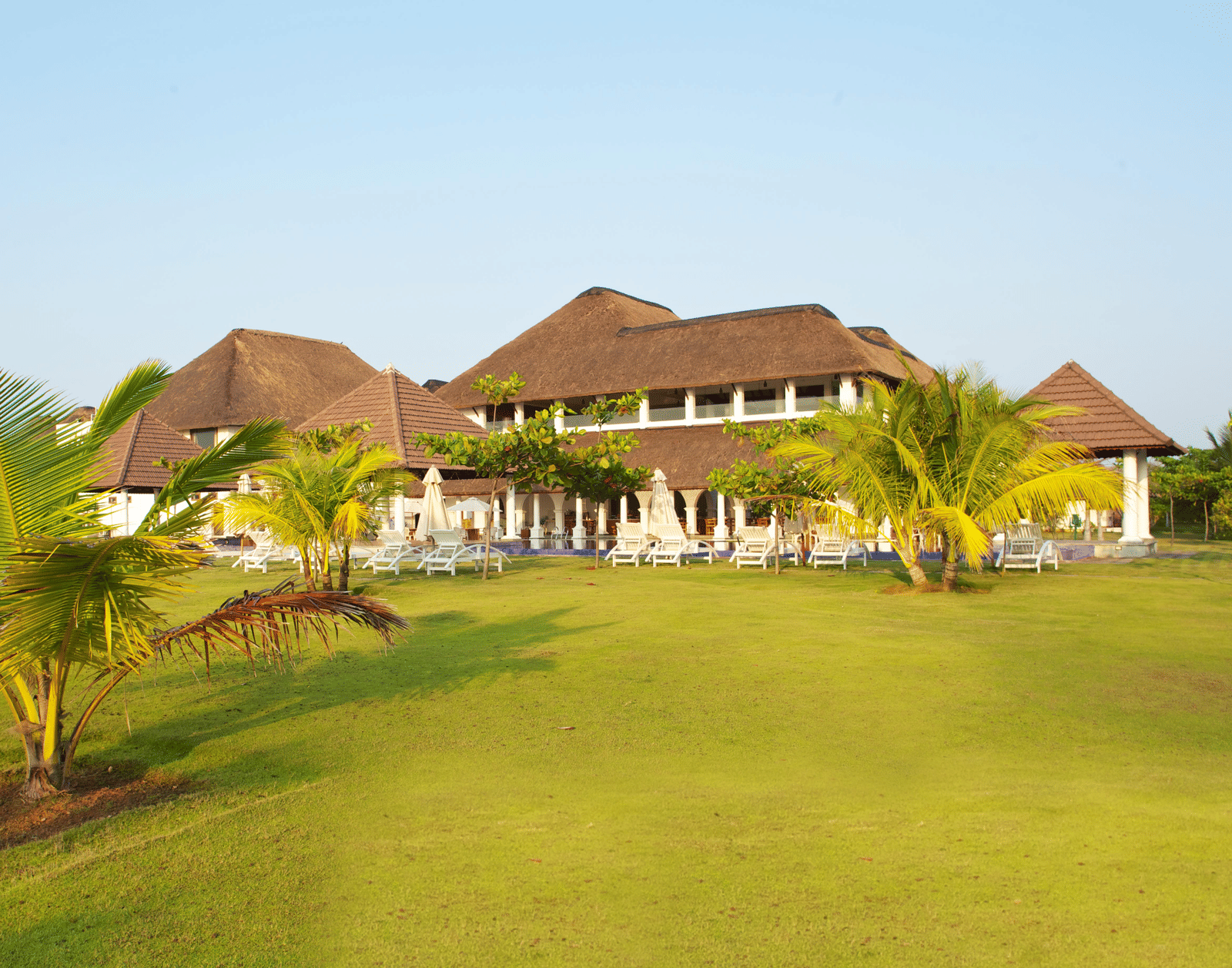 A wide green lawn with small palm trees situated in front of thatched-roof resort buildings | Le Pondy