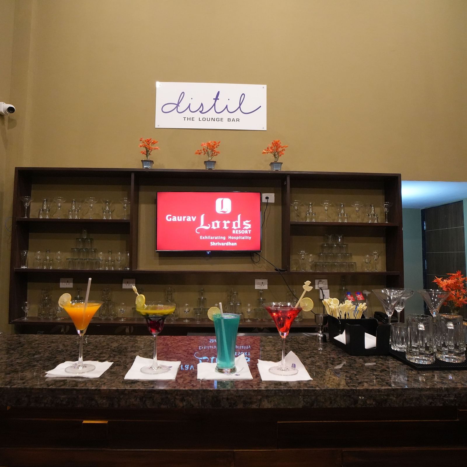 Bar counter with granite top showcasing colourful mocktails, cocktail glasses, napkins, and stirrers, backed by wooden shelves filled with glassware and minimal décor - Gaurav Lords Resort Shrivardhan