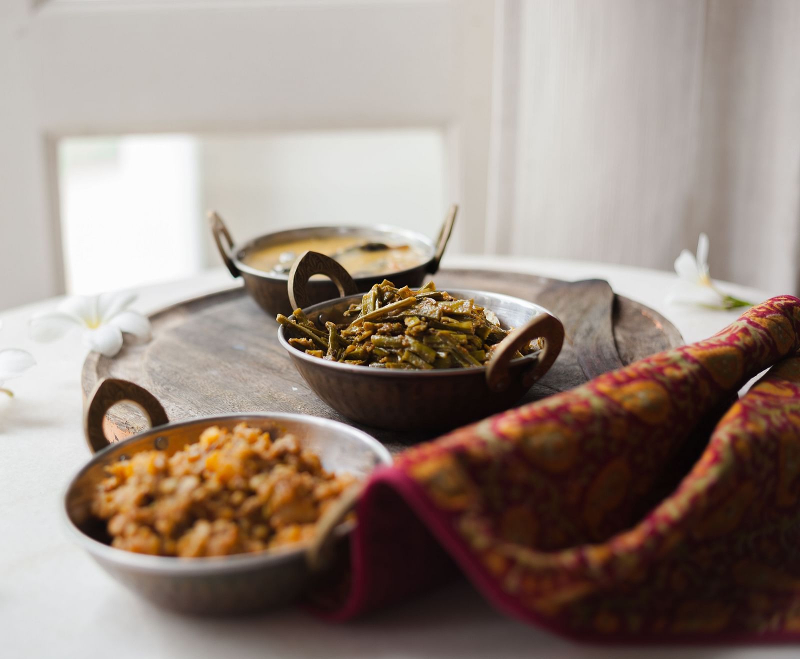 Three bowls of food on a tray, accompanied by a patterned cloth and flowers on a table- Shahpura Bagh.