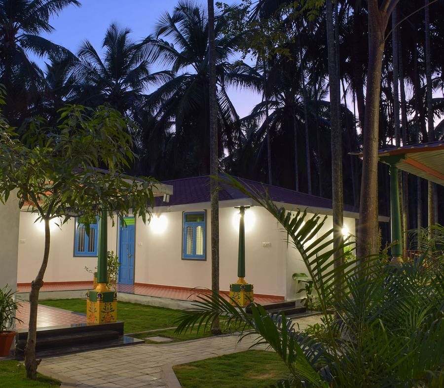 A white resort cottage with a red roof and blue window frames, surrounded by palm trees and green lawns - Ibex Resorts, Coimbatore (Kakarla)