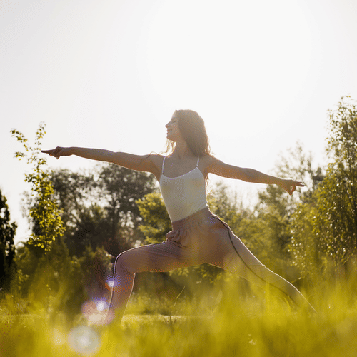 Woman practicing yoga outdoors in a grassy field, holding Warrior II pose with arms extended, surrounded by sunlight and trees