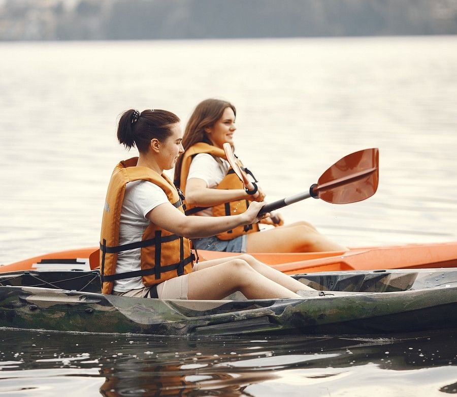 alt-text Two women in life jackets kayaking on a calm lake, surrounded by serene water and lush greenery.