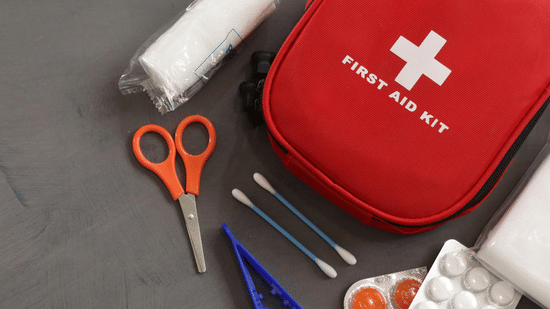 A red-coloured pouch reading 'First-aid Kit' surrounded by a pair of tweezers, Q-tips, a small scissor, and some medicines.