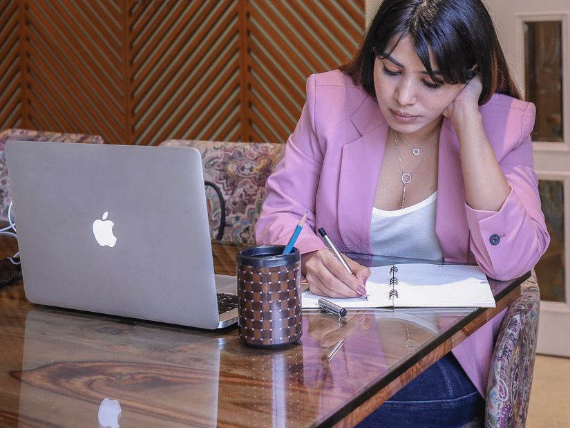 A woman in a pink blazer working on a laptop and writing in a notebook at Theory9 - Premium Service Apartments, Bandra, with water bottles and a coffee mug on the table.