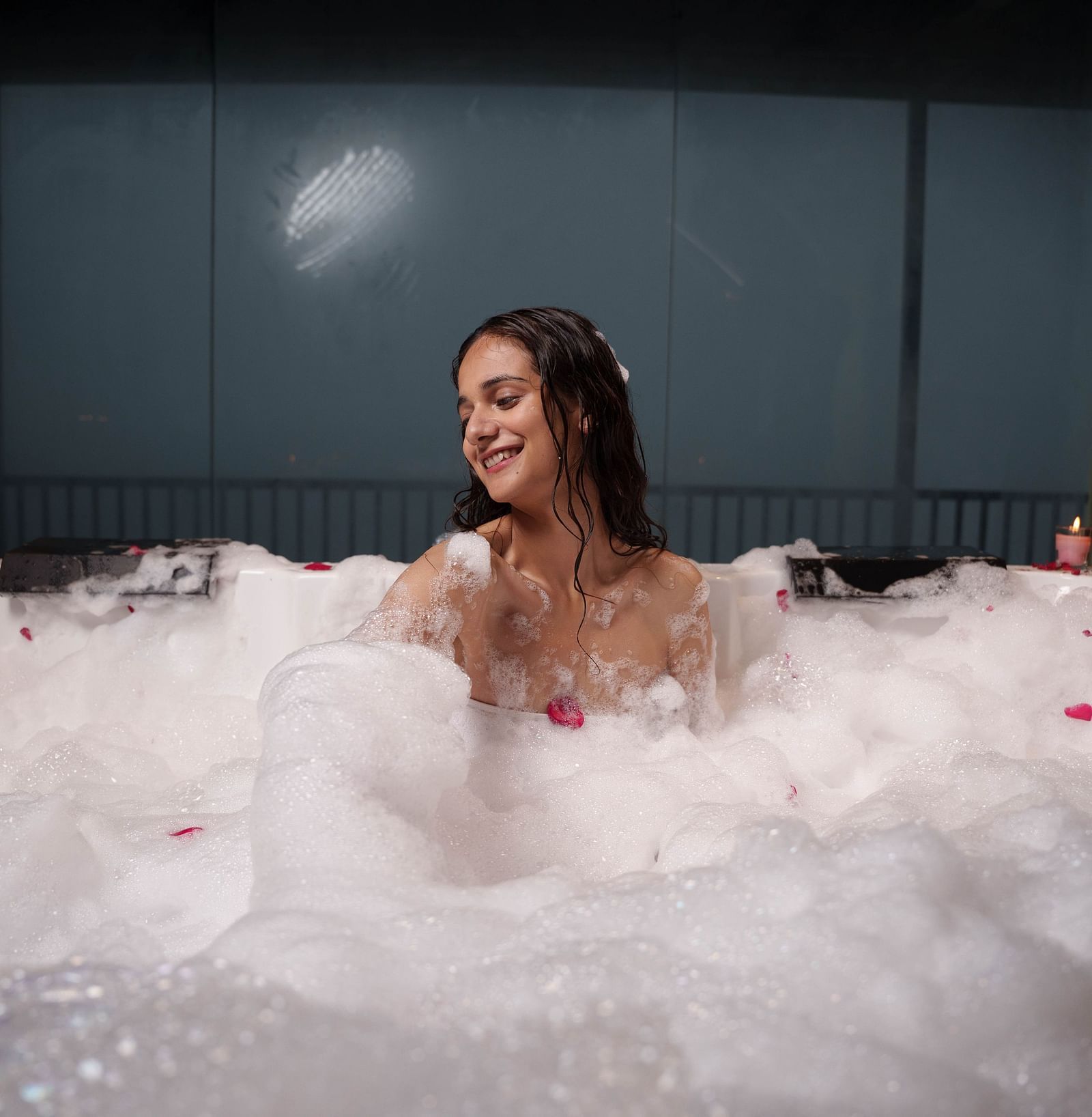 A woman is relaxing in a luxurious bubble bath, surrounded by white foam and rose petals, suggesting a spa experience at Summit Enigma Resort and Spa.
