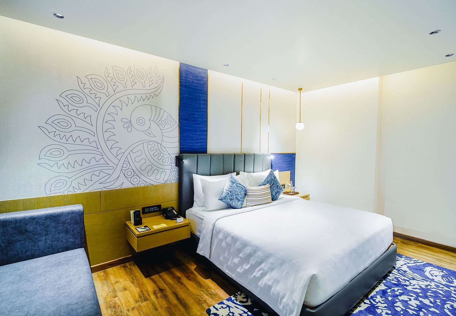 An image of our Deluxe Suite in Madurai with a peacock wall paper, a large bed, a night stand and a blue couch