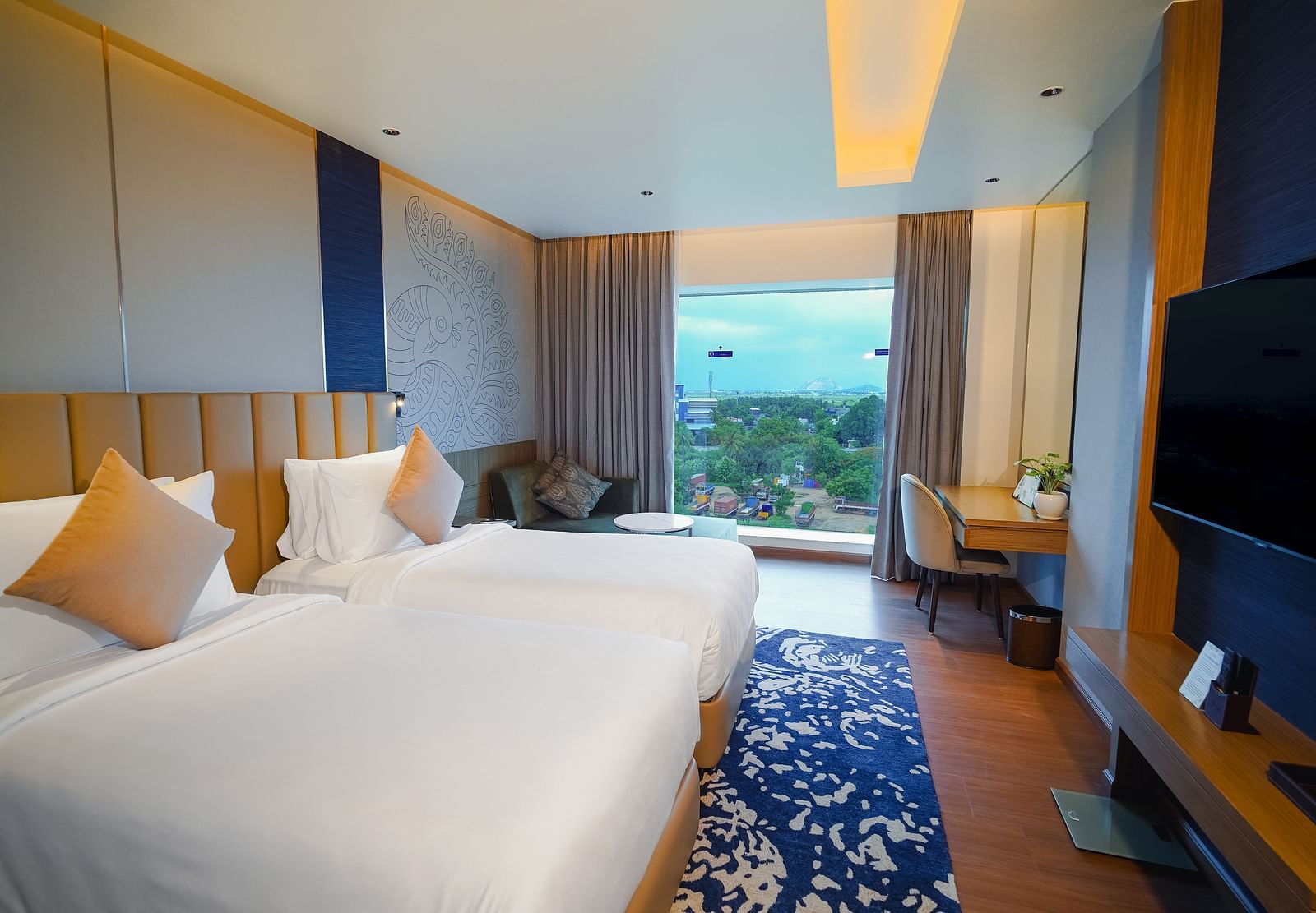 An image of a hotel room in Madurai with twin beds, a wall-mounted television, a work desk and a view of the hills