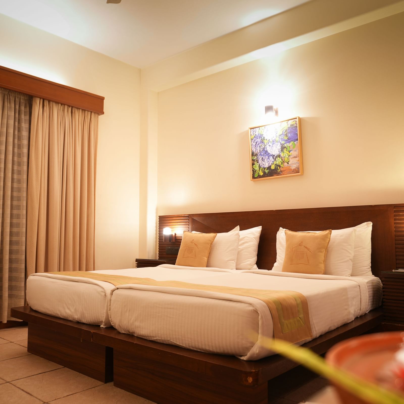 A warmly lit Superior Room at Vedic Village Spa Resort featuring a double bed with a wooden headrest, drapes over a large window, and a painting above the bed.