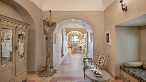 A hallway with arched doorways leading to other rooms, featuring a decorative cabinet, a sculpture, a table with chairs, and a built-in cushioned seating area in Tijara Fort-Palace - 19th Century, Alwar.