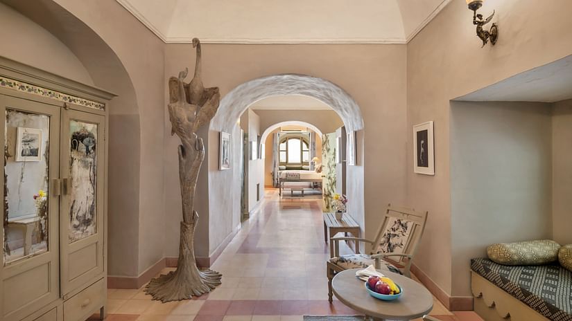 A hallway with arched doorways leading to other rooms, featuring a decorative cabinet, a sculpture, a table with chairs, and a built-in cushioned seating area in Tijara Fort-Palace - 19th Century, Alwar.