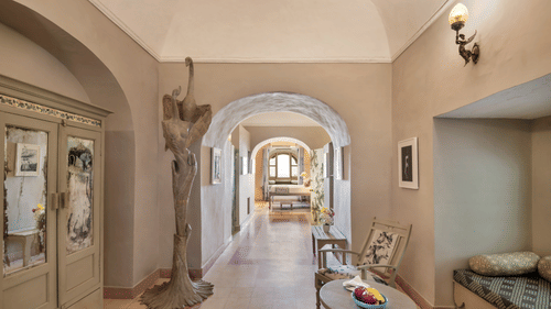 A hallway with arched doorways leading to other rooms, featuring a decorative cabinet, a sculpture, a table with chairs, and a built-in cushioned seating area in Tijara Fort-Palace - 19th Century, Alwar.