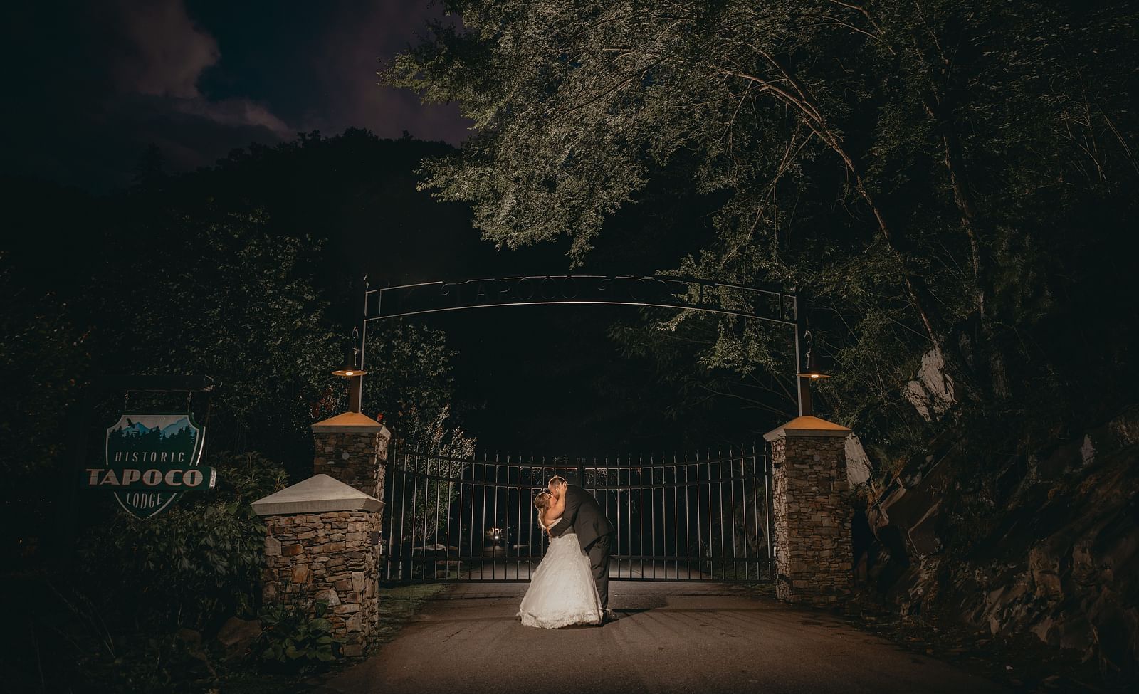 A newly wed couple kissing in front of a gate of the Historic Tapoco Lodge during the night time. The entrance also has trees on either side.