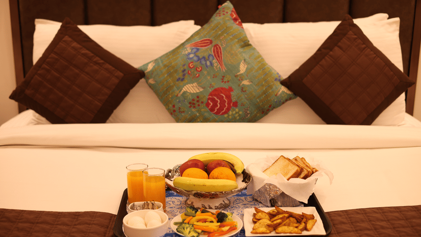 A centered shot of a hotel bed with colorful pillows and a breakfast tray with a variety of food and beverages.