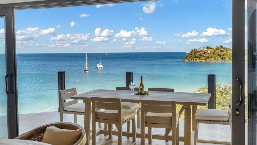 A dining area at Tamarind Hills Resort and Villas, Antigua with a wooden table and chairs positioned in front of floor-to-ceiling windows overlooking the sea and beach.