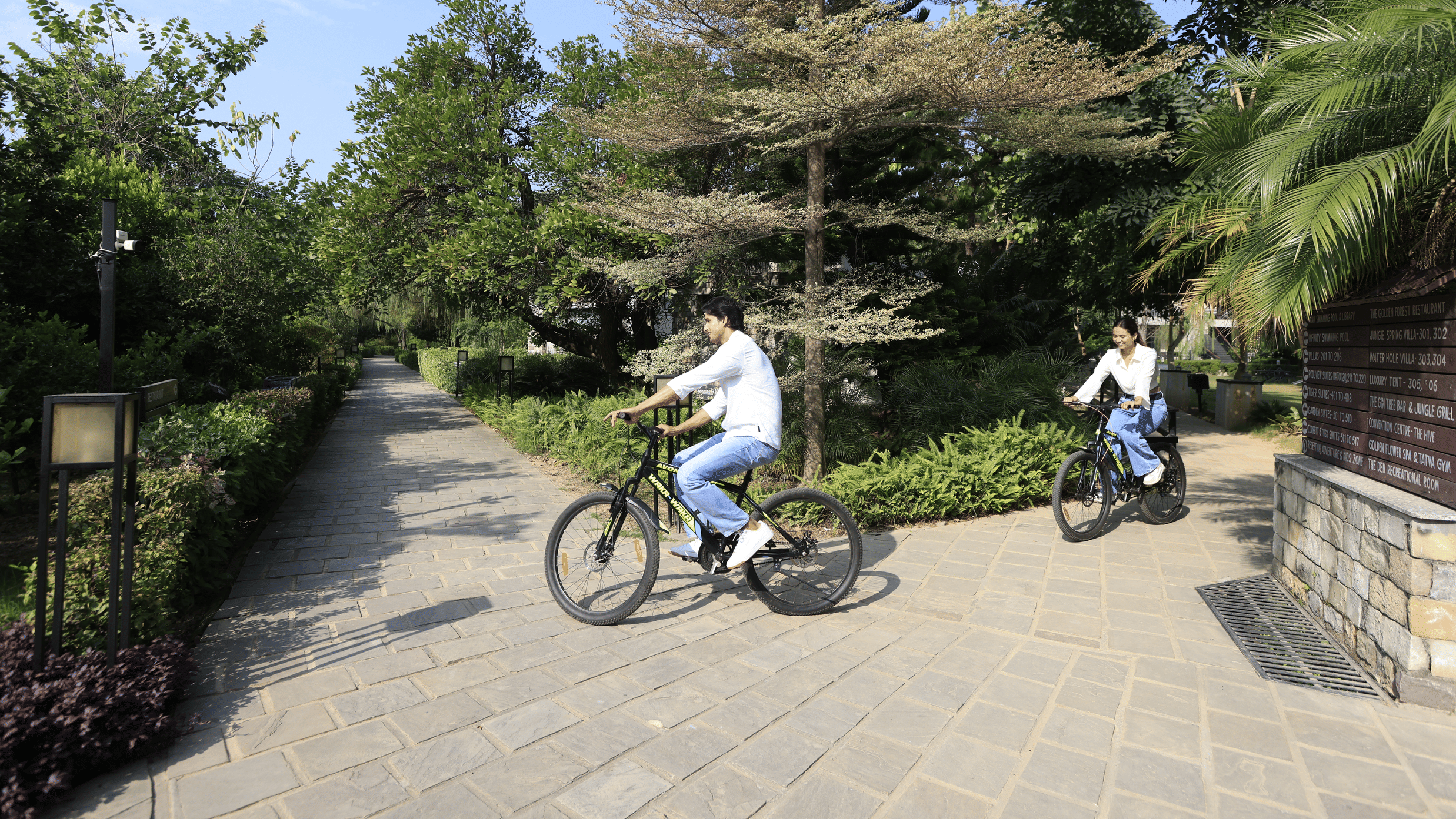 Two people riding bicycles on a paved path outdoors with trees and greenery at The Golden Tusk, Jim Corbett
