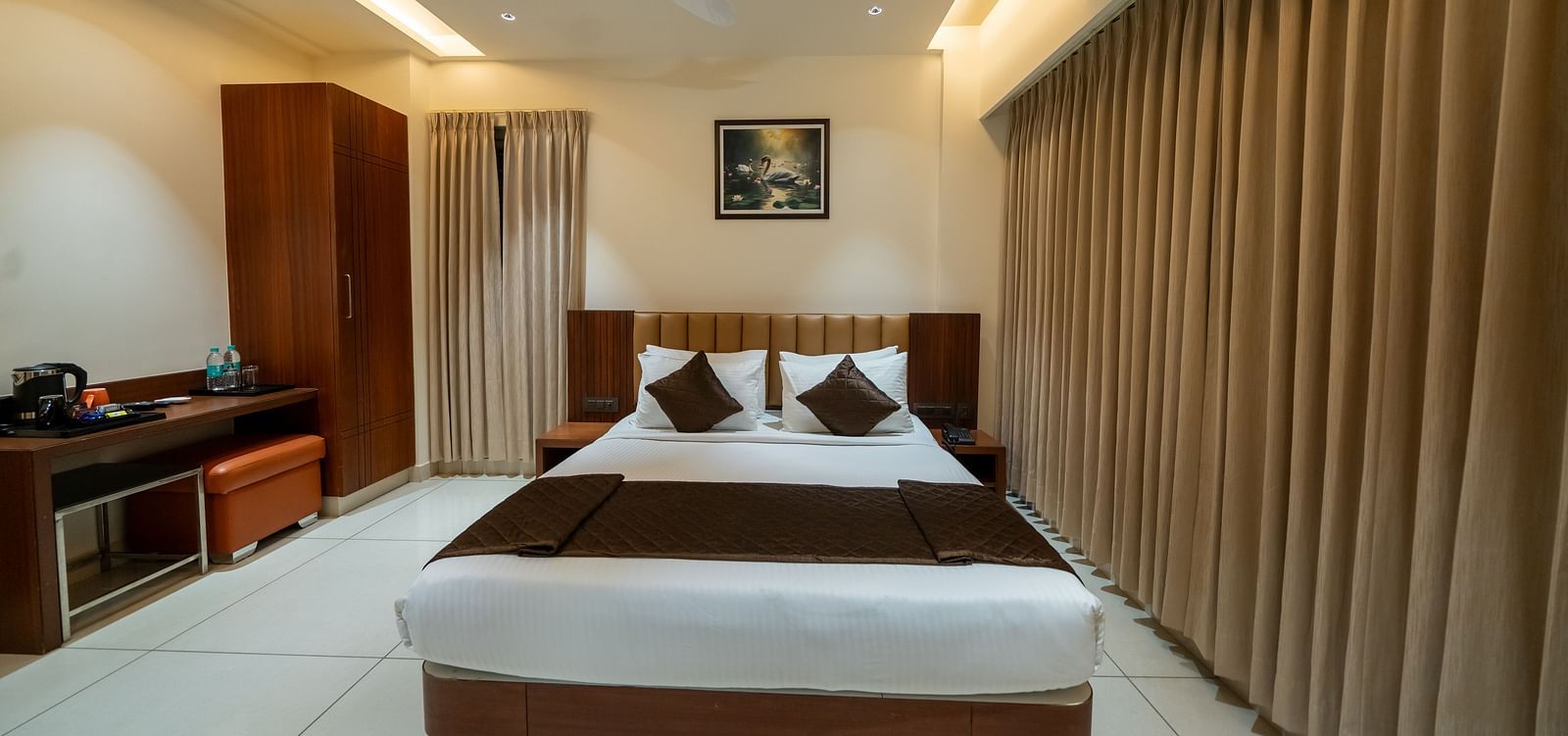 A front view of the bed at Hari Govinda's Express, Vijayanagar, with a photo frame above the headboard. To the left, a wooden cupboard stands beside a table with essentials, while on the right, an AC is mounted above the fully drawn curtains.