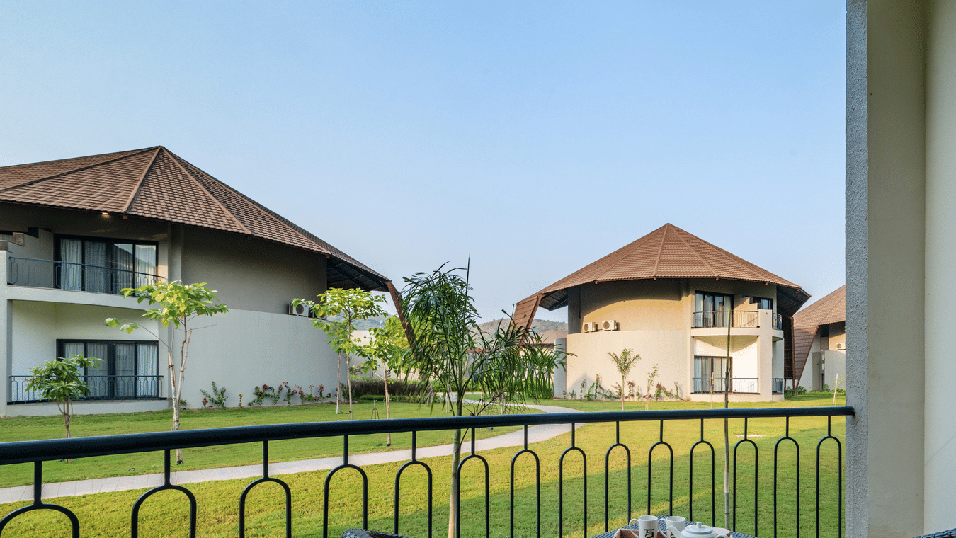 An image of a balcony with two wooden chairs and a tea table from where other villas and lawn can be seen at Ananta Spa and Resort, Ajabgarh