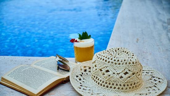 A glass of juice and a hat placed on a poolside lounge chair next to the swimming pool, with the water shimmering in the background.
