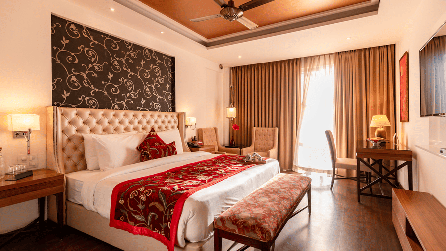 A bedroom with a king size bed, side tables, lamps, bench, curtains, and wall-mounted TV inside at Indraprastha Resort & Spa, McLeod Ganj.