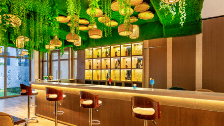 Wide view of the bar at Lords Inn Sangam, Bhopal with a curved wooden counter, red stools, bottle display shelves, and green ceiling decor.