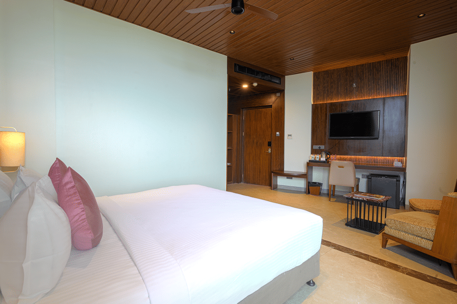 Hotel bedroom with a king size bed, pink accent cushions, bedside lamp, television and wooden doorway leading to the entrance area.