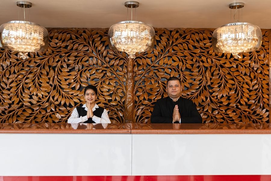Reception area of Pride Premier Dehradun with staff welcoming guests in traditional Indian style, set against an intricately carved wooden backdrop and elegant pendant lighting.