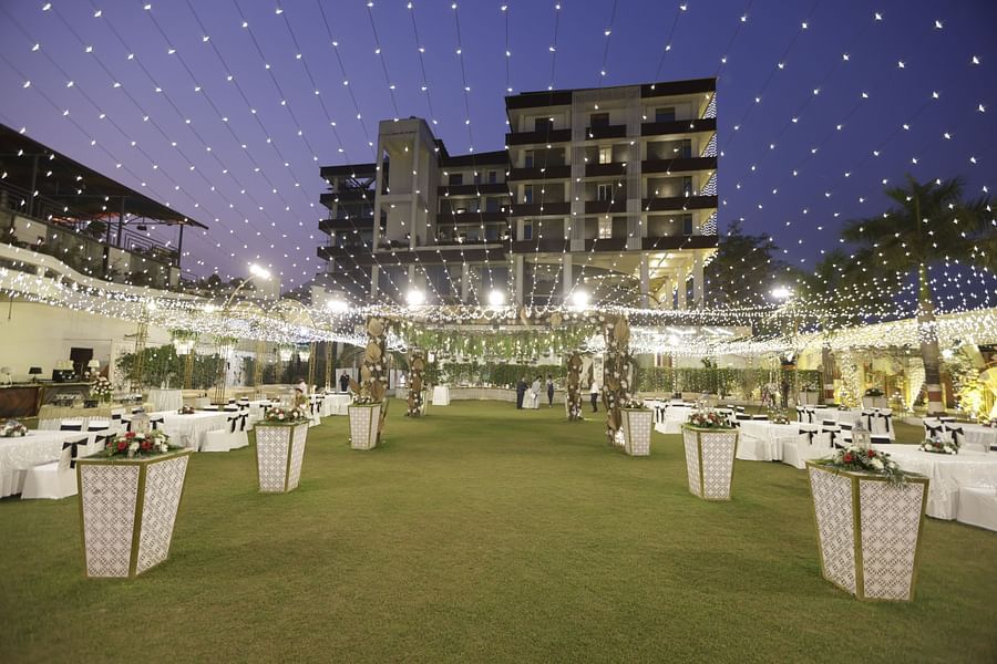 Pride Premier Dehradun featuring outdoor wedding setup on a lush lawn with hanging string lights, floral decor, banquet seating, and a modern building backdrop at dusk.