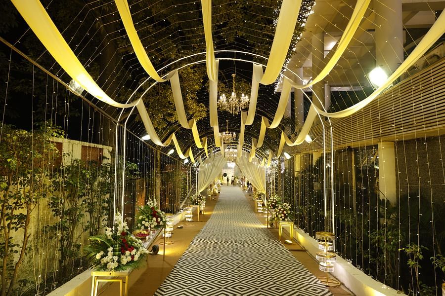 A warmly lit wedding walkway with floral arrangements, chandeliers, draped fabric, and patterned carpet under a canopy of fairy lights at Pride Premier Dehradun