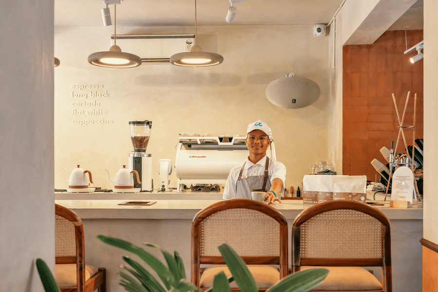 A staff member in white uniform and cap standing behind a clean cafe counter at The Bakehouse with coffee equipment wooden cabinetry rattan chairs and warm overhead lighting in a bright setting.