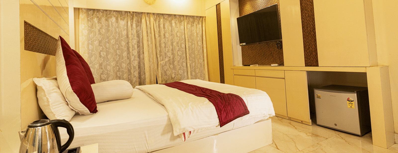 A bedroom with a king size bed, a bedside table with an electric kettle, a television, a mini fridge, and curtains at Hotel Seagull Digha.