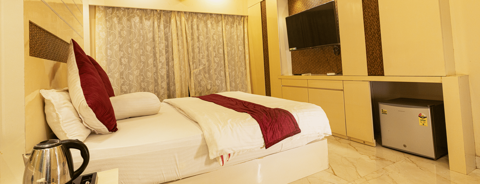 A bedroom with a king size bed, a bedside table with an electric kettle, a television, a mini fridge, and curtains at Hotel Seagull Digha.