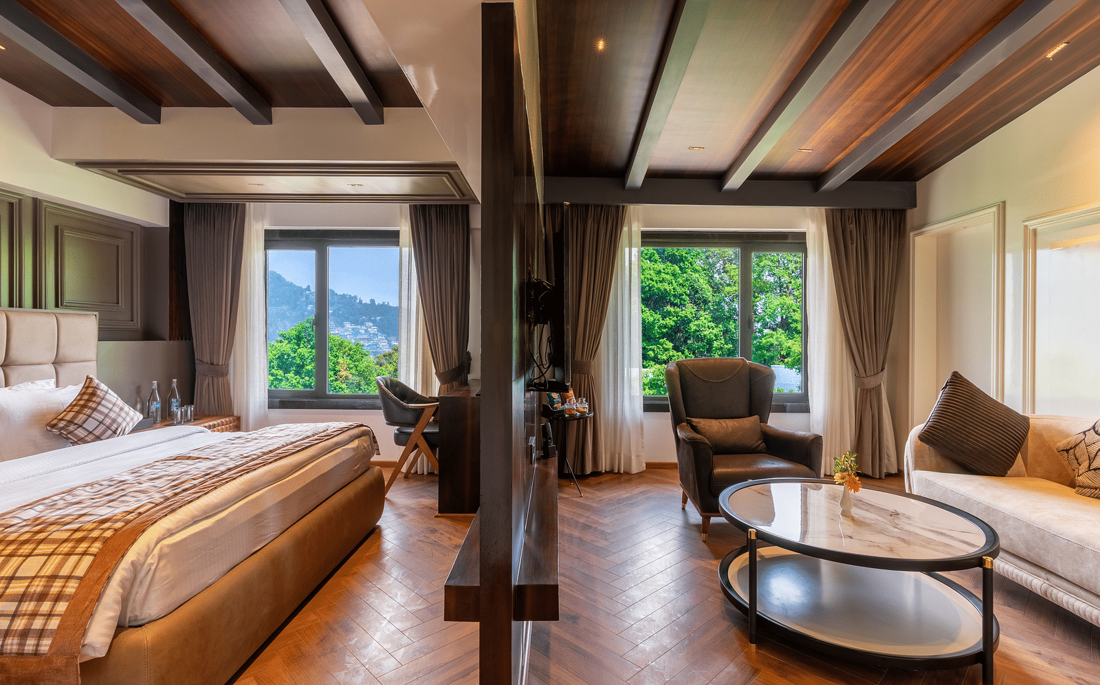 An image of a bedroom with partition, on one side is a king size bed, a chair and table and on the other side is a seating area with sofa set and a centre table