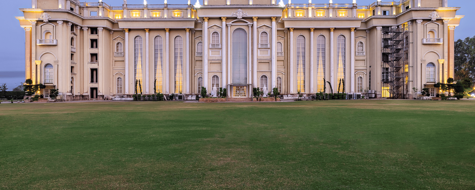 Vast, ornate, pale-coloured building with a central dome and grand architecture, illuminated against a twilight sky, set on a lawn at Hotel Hukam's Lalit Mahal.