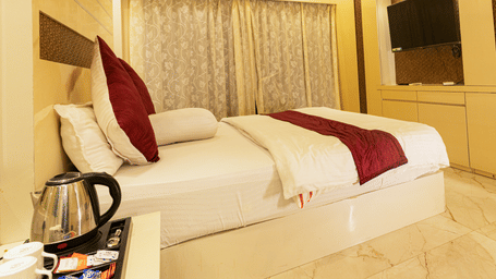 A bedroom with a king size bed, a bedside table with an electric kettle, a television, a mini fridge, and curtains at Hotel Seagull Digha.