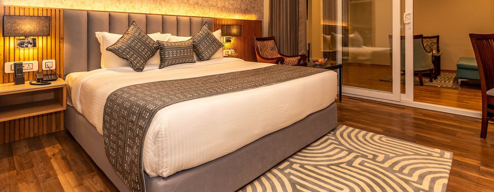 An image of a Family Room with a bed, bed side table, lamps, rug, and slider glass door leading to the other areas of the room - The Belvedere Himalayan Retreat, McLeodganj