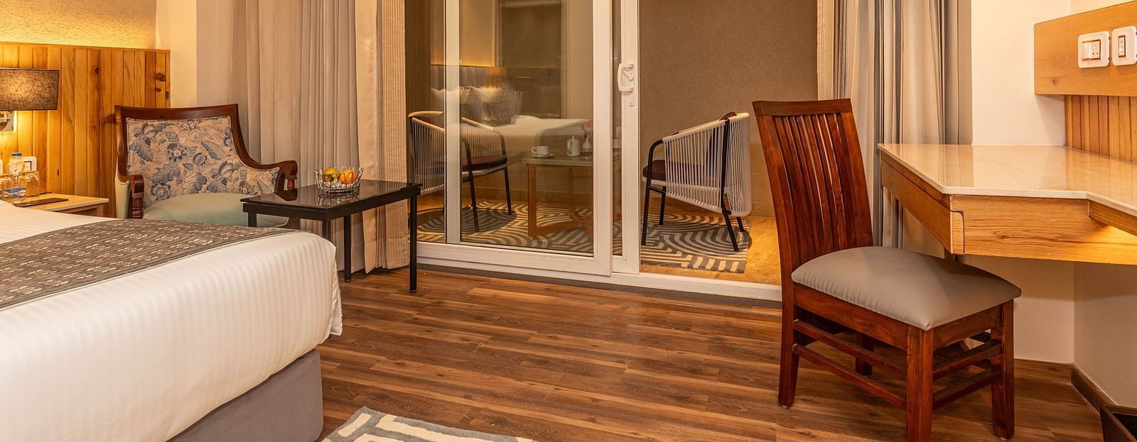 An image of Deluxe Room with a bed, work desk and chair, sofa and table, and an expansive glass door leading to the balcony - The Belvedere Himalayan Retreat, McLeodganj