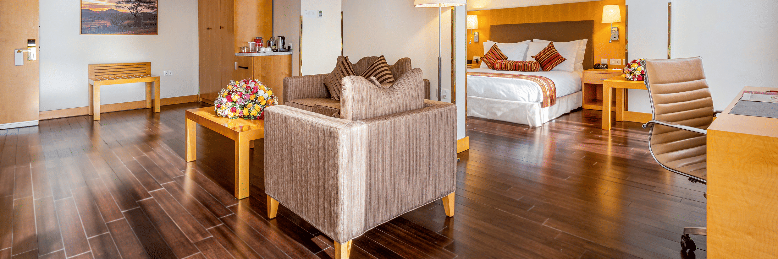 A suite with a bed, seating area, desk, chairs, tables, lamps, wooden floor, wall art, wardrobe, and a tea and coffee setup at Ramada by Wyndham Addis, Addis Ababa.