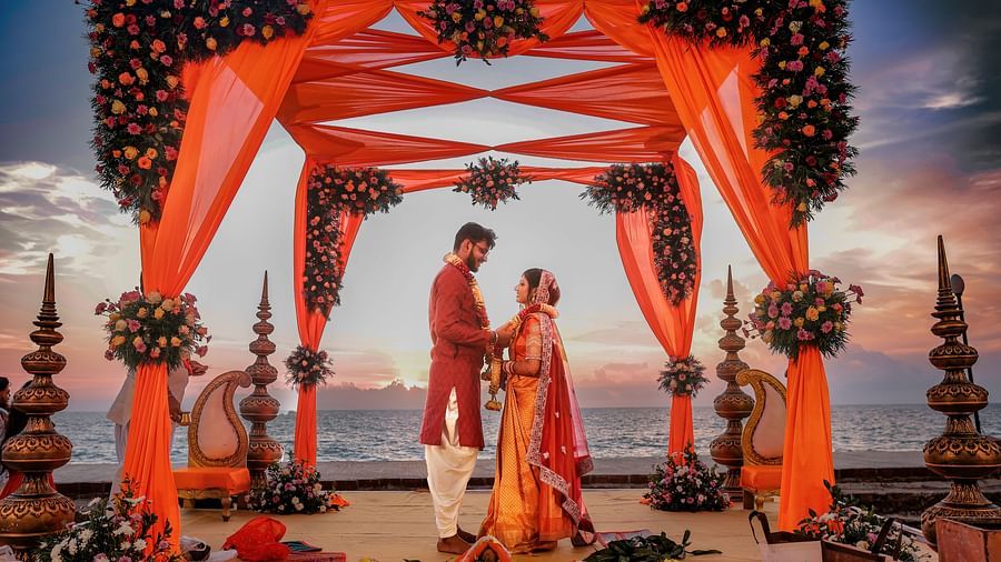 An image of a couple standing together in the wedding mandap by the beach at sunset, with a beautifully decorated floral design as they exchange vows