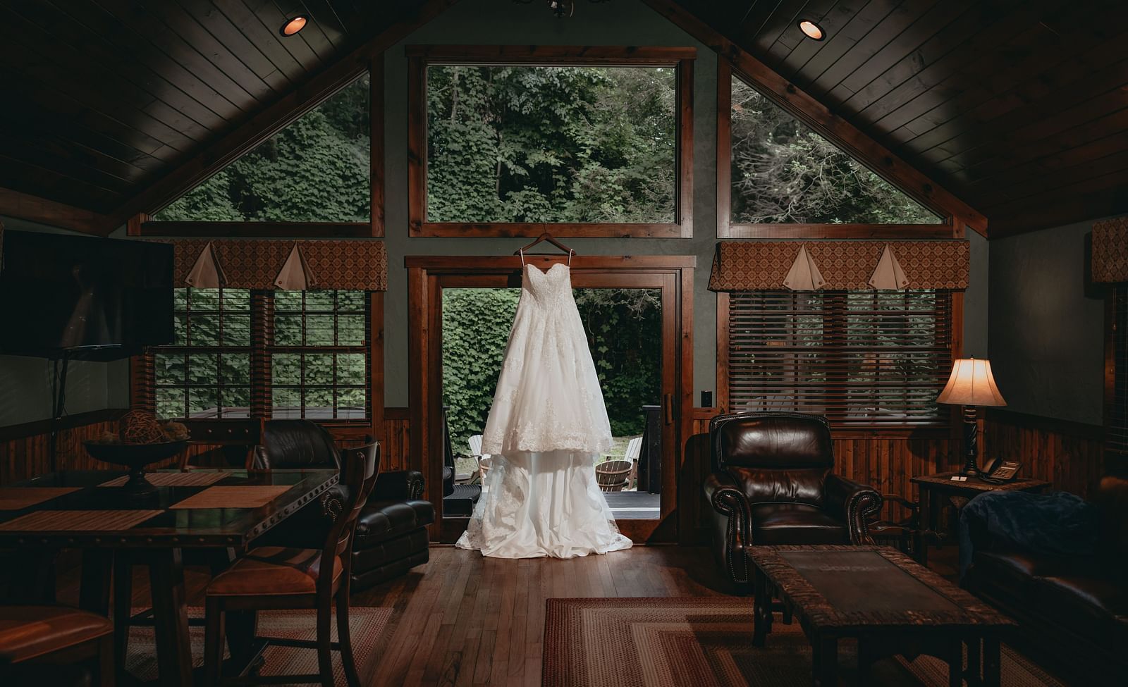 A wedding gown hanged at the entrance of a wooden cottage at the Historic Tapoco Lodge. The room also features a lit lamp at the corner, sofa chairs, dining table paired with four chairs.