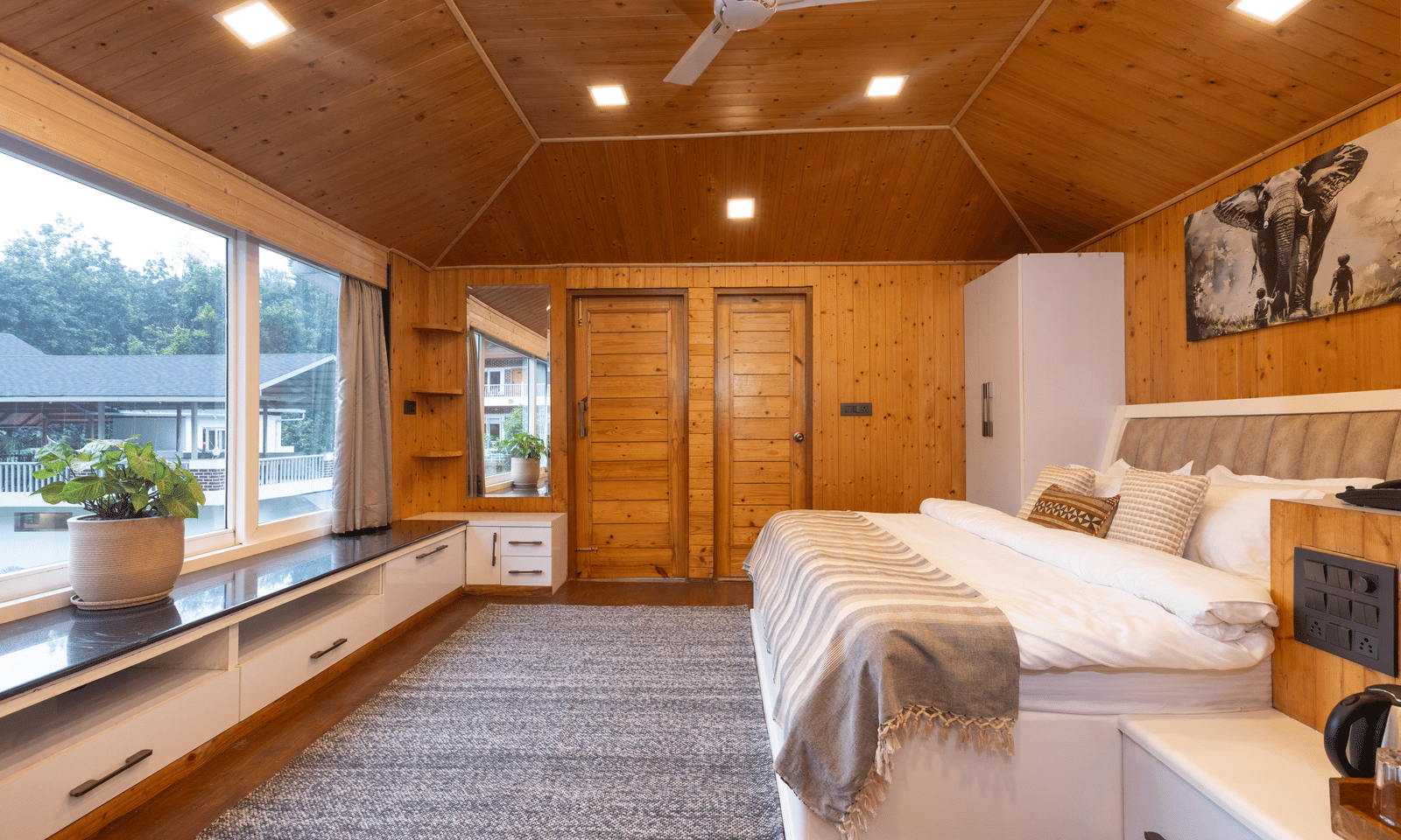 A spacious wooden bedroom has wide windows and a large bed with a view of the outdoors at Megha Resort & Villas, Dehradun.