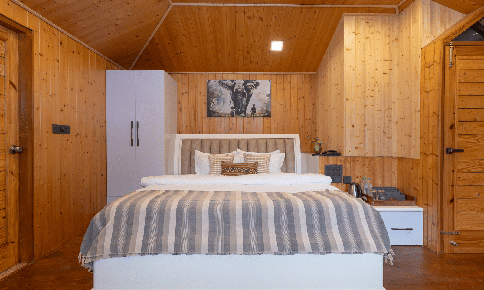 A cosy bedroom features wooden walls and a double bed with a striped neutral-toned coverlet at Megha Resort & Villas, Dehradun.