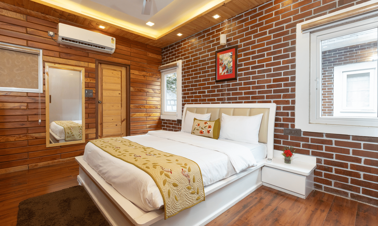 A well-lit bedroom showcasing the red brick and wooden wall behind the large, comfortable bed at Megha Resort & Villas, Dehradun.