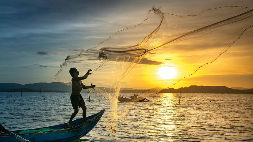 An image of a man throwing a fishing net in the sea with the backdrop of the setting sun