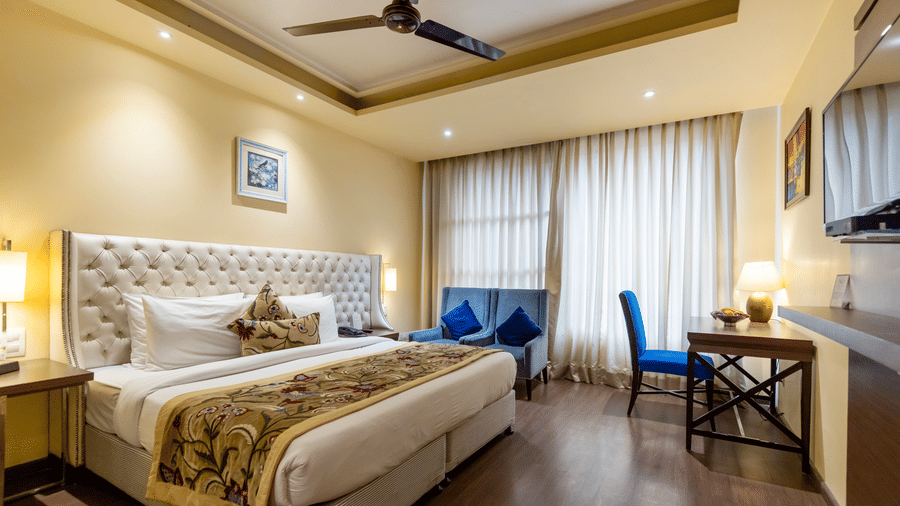 A bedroom with a king size bed, side tables, work desk, chair, lamps, curtains, and wall-mounted TV at Indraprastha Resort & Spa, McLeod Ganj.