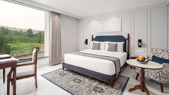 An image of our hotel room in Haridwar featuring a large bed, an upholstered sofa, a round table, and a scenic view - MJ Sarovar Portico, Haridwar