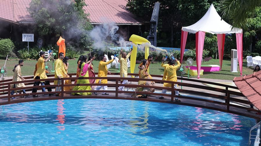 An image of people in colourful traditional attire, walking over a wooden bridge above a pool while holding smoke guns releasing coloured smoke