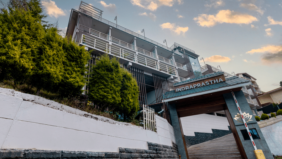 An exterior view of Indraprastha Resort & Spa, McLeod Ganj showing building facade, balconies, entrance gate, and surrounding greenery.