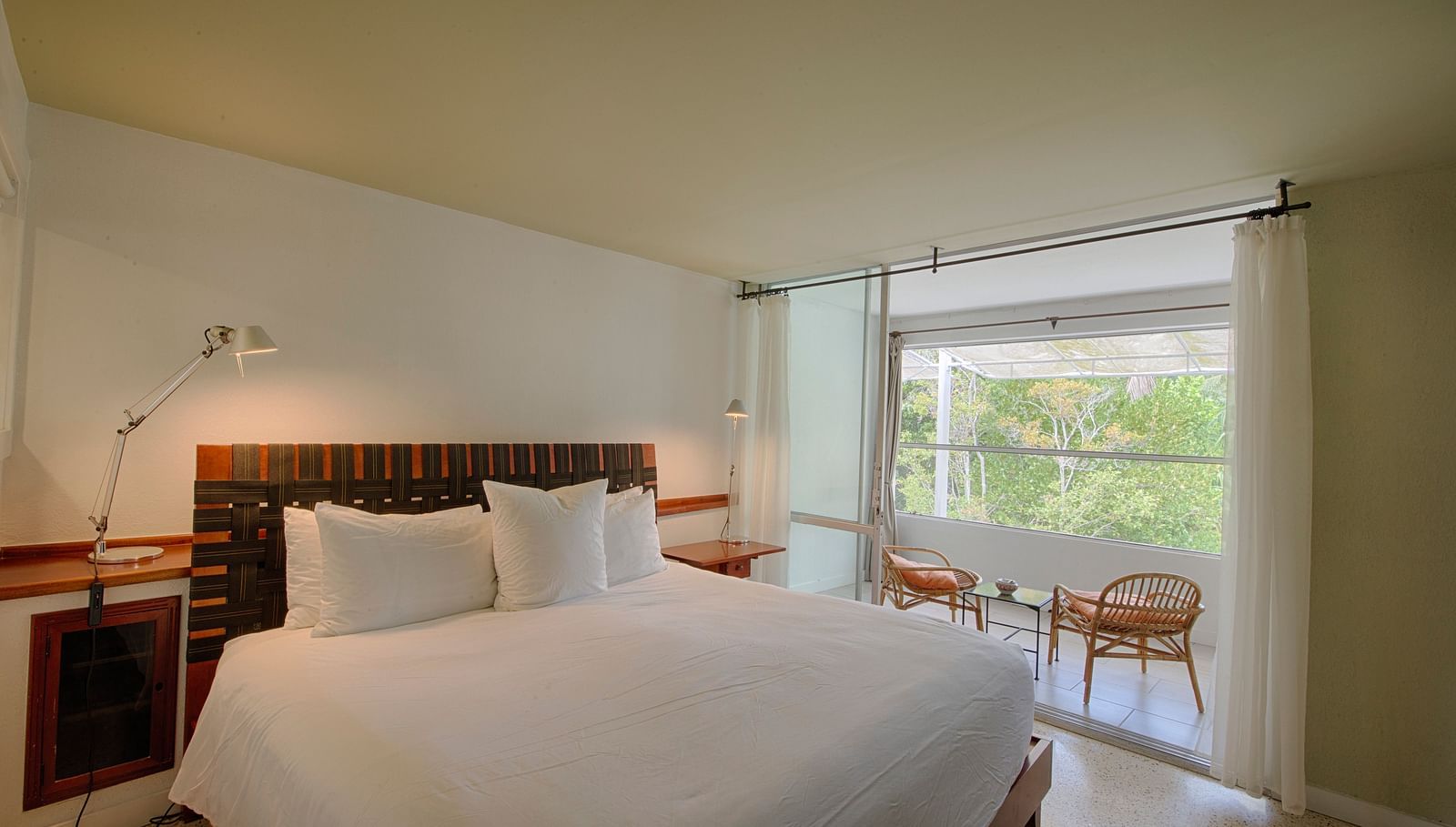 Bed with white linens at Casa Morada, facing a window with a garden view and two chairs.